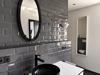 Ein modernes Badezimmer mit grauen Wandfliesen und einem runden Spiegel. Die Einrichtung ist minimalistisch mit einem schwarzen Waschbecken und einer eleganten Toilette im Hintergrund.