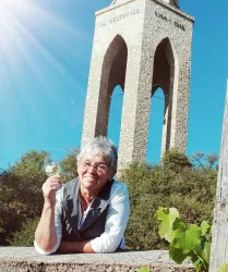 Eine lächelnde Frau hält ein Glas in der Hand, während sie vor einem hohen Steinbau steht. Im Hintergrund ist ein klarer blauer Himmel und die Sonne scheint.
