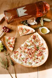 Eine köstliche Pizza mit Schinken und Gemüse liegt auf einem Holztisch. Daneben steht eine Flasche Roséwein und frische Zutaten.