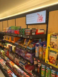 Ein Regal in einem Geschäft mit verschiedenen Süßigkeiten und Snacks. Oben an der Wand hängt ein Bildschirm mit einer Anzeige.