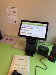 Ein moderner Kiosk mit einem Tablet, der eine Willkommensnachricht und verschiedene Produkte anzeigt. Auf dem Tisch liegen Informationsmaterialien und ein Kartenlesegerät.