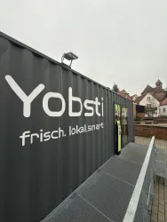 Ein moderner Container mit der Beschriftung "Yobsti" und dem Slogan "frisch. lokal. smart.". Im Hintergrund sind einige historische Gebäude und ein grauer Himmel zu sehen.
