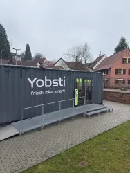 Ein moderner Verkaufscontainer mit der Aufschrift "Yobsti" steht in einer ruhigen Umgebung. Der Container bietet frische, lokale Produkte an und hat einen Rampenzugang.