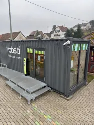 Ein moderner Laden in einem Versandcontainer, umgeben von einer gepflasterten Fläche. Die Fenster sind offen und der Eingangsbereich ist einladend gestaltet.