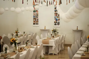 Ein festlich dekorierter Veranstaltungsraum mit langen Tischen und weißen Stühlen. Die Wände sind mit Fotos und Dekorationen geschmückt, und es gibt Blumenarrangements auf den Tischen.