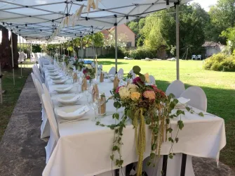 Ein festlich gedeckter Tisch unter einem Zelt, umgeben von grünem Rasen. Blumenarrangements schmücken die Tische in einer einladenden Umgebung.