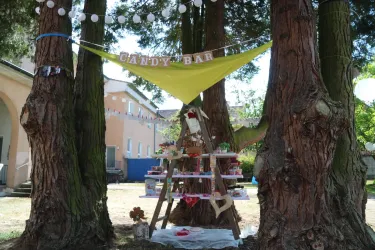 Eine süße Candy-Bar, dekoriert mit bunten Süßigkeiten und einer Wimpelgirlande. Sie ist zwischen zwei großen Bäumen aufgestellt und sorgt für eine einladende Atmosphäre.