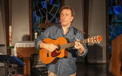Ein Mann spielt Gitarre und singt in einem Raum mit bunten Fenstern. Es ist eine gemütliche Atmosphäre mit einem Tisch auf der linken Seite.