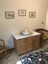 Ein stilvoll eingerichteter Raum mit einem Holzschrank und einem weißen Krug auf einem Tisch. An der Wand hängen drei Bilder, und ein Stuhl sowie ein Korbsessel ergänzen die Einrichtung.