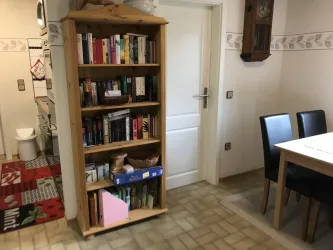 Ein gemütlicher Raum mit einem Bücherregal voller Bücher. An der Wand sind ein Tisch und schwarze Stühle zu sehen.