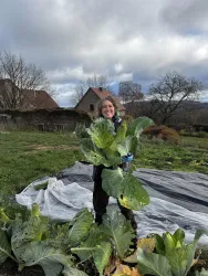 Eine Person steht auf einem Feld und hält große Kohlköpfe in den Händen. Im Hintergrund sind Bäume und ein Haus zu sehen.