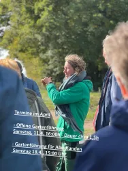 Eine Gruppe von Menschen hört einer Frau in einem grünen Mantel zu, die spricht. Im Hintergrund sind Bäume und die Natur zu sehen.