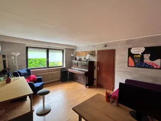 Ein helles Wohnzimmer mit einem Klavier, blauen Sessel und einem Tisch. An der Wand hängt ein buntes Kunstwerk.