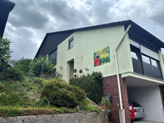 Ein zweigeschossiges Haus mit einem begrünten Hang und einem sichtbaren Auto in der Garage. Der Himmel ist bewölkt und es gibt eine bunte Wandmalerei an der Seite des Hauses.