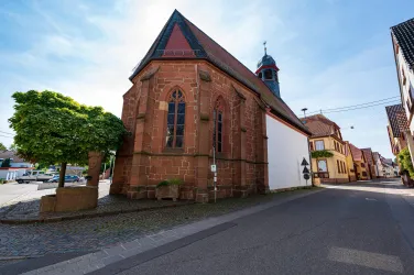 Eine alte Kirche aus roten Ziegeln steht an einer Straße. Im Hintergrund sind weitere Gebäude und Bäume sichtbar.