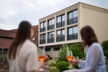 Ein modernes Gebäude mit mehreren Fenstern und Balkonen. Zwei Personen genießen Drinks im Freien, umgeben von Gartenpflanzen.