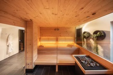 Eine moderne Sauna mit Holzverkleidung und sanfter Beleuchtung. Rechts befindet sich ein Steingrill und an der Wand hängen Pflanzenarrangements.