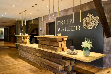 Der Empfangsbereich eines Hotels mit einem modern gestalteten Holztresen. Hinter dem Empfang steht ein elegantes Schild mit dem Namen "Fritz Walter".
