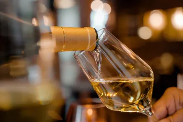 Ein Glas Wein wird aus einer Flasche eingegossen. Die Flüssigkeit hat eine goldene Farbe und wird in einem elegant gestalteten Glas serviert.