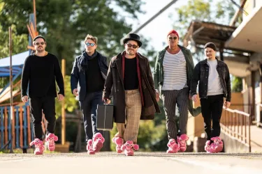 Eine Gruppe von fünf Männern läuft selbstbewusst auf einem Weg. Sie tragen ungewöhnliche pinkfarbene Hausschuhe und unterschiedliche stylische Outfits.