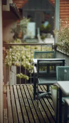 Ein kleiner Balkon mit Holzfußboden und modernen Möbeln. Im Hintergrund sind grüne Pflanzen zu sehen.