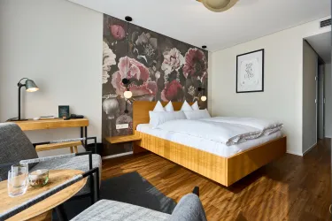 Ein modernes Hotelzimmer mit einem Doppelbett und einer blumigen Wandtapete. Es gibt einen gemütlichen Leseplatz und viel natürliches Licht.