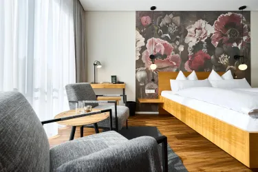 Ein modernes Zimmer mit einem großen Bett und einem dekorativen Blumenhintergrund. Gemütliche Sessel und ein Tisch bieten einen einladenden Platz zum Entspannen.