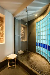 Eine moderne Dusche mit einer stilvollen Glaswand und einem farbigen Lichtpaneel. Der Boden ist mit runden Steinen gestaltet, und ein kleiner Hocker steht bereit.