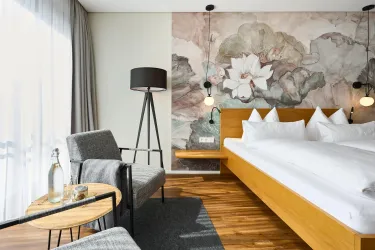 Ein modernes Hotelzimmer mit einem großen Bett und eleganten Kissen. Die Wände sind mit floralen Motiven gestaltet, und es gibt gemütliche Möbel sowie viel Tageslicht.