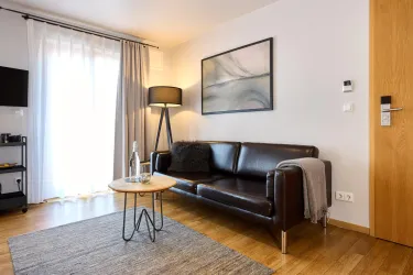 Ein modernes Hotelzimmer mit einem braunen Sofa, einem Holztisch und einem Teppich. Große Fenster sorgen für viel Tageslicht.