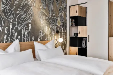 Ein modernes Hotelzimmer mit einem großen Bett und einer dekorativen Wand mit Pflanzenmotiven. Der Raum ist hell und einladend, mit einem Holzregal zur Aufbewahrung.