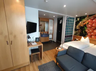 Ein modernes Hotelzimmer mit Holzfußboden, einem gemütlichen Bett und einem grauen Sofa. An der Wand gibt es eine Fototapete von Trauben, und es gibt einen TV sowie einen Schreibtisch.