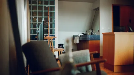 Ein gemütlicher Raum mit einem Holzstuhl im Vordergrund. Im Hintergrund sind eine Badecke und Glasbausteine der Dusche sichtbar.