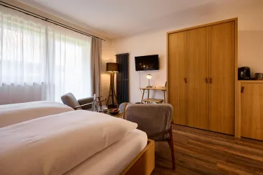 Ein modernes Zimmer mit zwei Einzelbetten und Holzfußboden. Die Einrichtung umfasst einen Sessel, einen Nachttisch und einen Fernseher an der Wand.