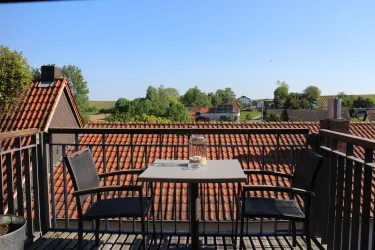 Ein Balkon mit einem Tisch und zwei Stühlen. Im Hintergrund sind grüne Felder und Häuser zu sehen unter einem klaren blauen Himmel.