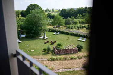 Eine grüne Gartenlandschaft mit Liegen und einem kleinen Sitzbereich aus Holz. Im Hintergrund sieht man Bäume und eine weite, offene Landschaft.