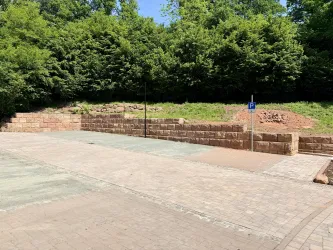 Ein Parkplatz mit Pflastersteinen und einer begrünten Wand aus Natursteinen. Im Hintergrund sind Bäume zu sehen.