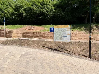 Ein Parkplatz mit einem Informationsschild und einer gepflasterten Straße. Im Hintergrund sind grüne Bäume sichtbar.