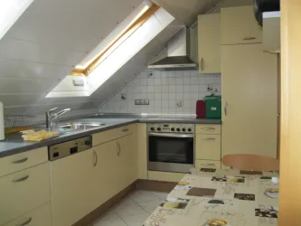 Eine moderne Küche mit hellen Schränken und einem Schrägdach. Ein Tisch mit Tischdecke steht im Raum und Fenster sorgt für Tageslicht.