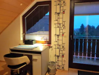Ein gemütliches Zimmer mit Schreibtisch und Stuhl. Durch das Fenster blickt man auf einen Balkon und die angrenzende Natur.
