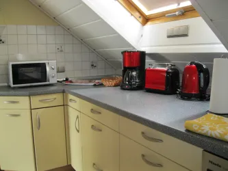 Eine moderne Küche mit gelben Schränken und einem hellen Tisch. Auf der Arbeitsplatte stehen eine Mikrowelle, eine Kaffeemaschine und ein Wasserkocher in auffälligem Rot.