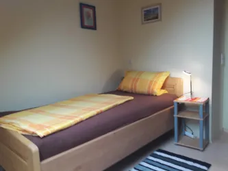 Ein einfaches Schlafzimmer mit einem Einzelbett und bunten Kissen. Neben dem Bett steht ein kleiner Nachttisch mit einer Lampe.