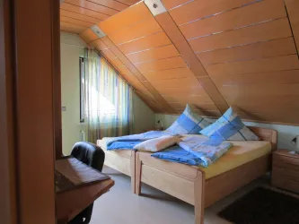Ein gemütliches Schlafzimmer mit einem Dachschrägen und zwei Betten. Die Wände sind hell, und es gibt Fenster mit bunten Vorhängen.