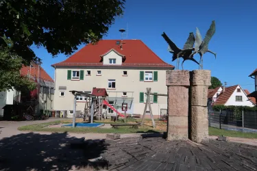 Ein Spielplatz mit verschiedenen Spielgeräten und einer Skulptur befindet sich vor einem Gebäude mit rotem Dach. Die Umgebung ist sonnenbeschienen und grün.
