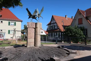 Ein Brunnen mit einer Skulptur von Vögeln steht im Mittelpunkt eines Platzes. Umgeben ist er von traditionellen Gebäuden und grünen Bäumen.