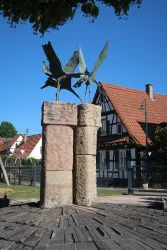 Eine Skulptur von zwei fliegenden Vögeln auf zwei Säulen aus Stein. Im Hintergrund steht ein traditionelles, zweigeschossiges Fachwerkhaus.