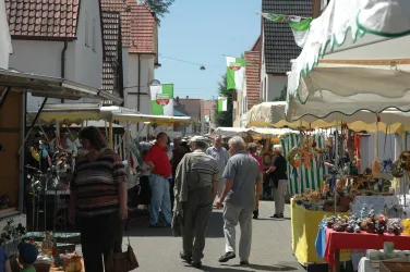 Ein belebter Markt mit Ständen und Menschen, die umherlaufen. Die Stände bieten verschiedene Waren unter Sonnenschirmen an.