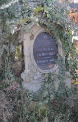 Ein altes Grabmal, umgeben von Efeu. Es zeigt den Namen und die Lebensdaten einer Person.