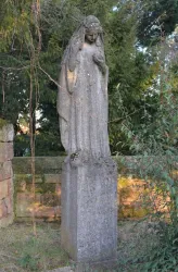 Eine Steinstatue einer weiblichen Figur steht auf einem Sockel. Die Statue ist von Bäumen und Pflanzen umgeben.