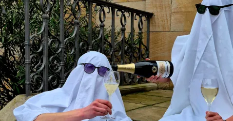 Zwei Personen in weißen Laken tragen Sonnenbrillen und genießen Champagner. Eine Person gießt Champagner in ein Glas, während die andere entspannt sitzt.